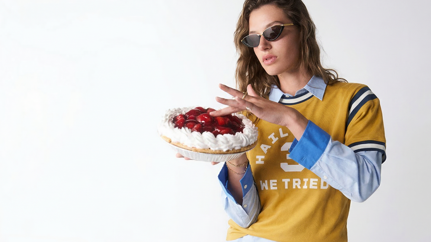 Woman holding a cake with strawberries, wearing sunglasses and a yellow and blue jacket.